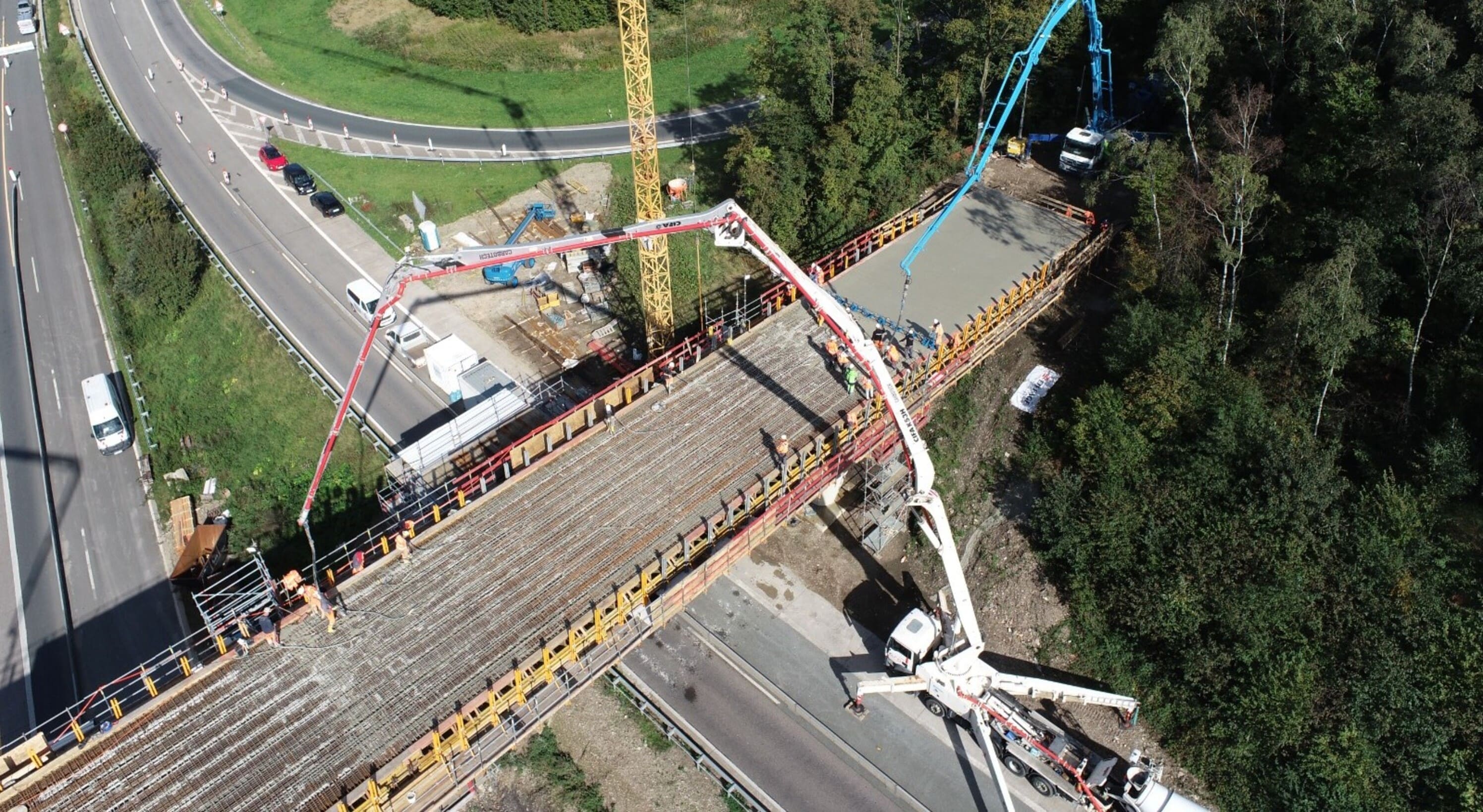 Luftaufnahme einer Brückenbaustelle: Zwei große Betonpumpen mit roten und blauen Auslegern verteilen frischen Beton auf einer Brücke. Die Brücke verläuft über eine Straße und ist von dichtem Wald umgeben. Auf der Brücke sind Arbeiter bei der Arbeit zu sehen, während unten Fahrzeuge auf der Straße fahren.

                                      